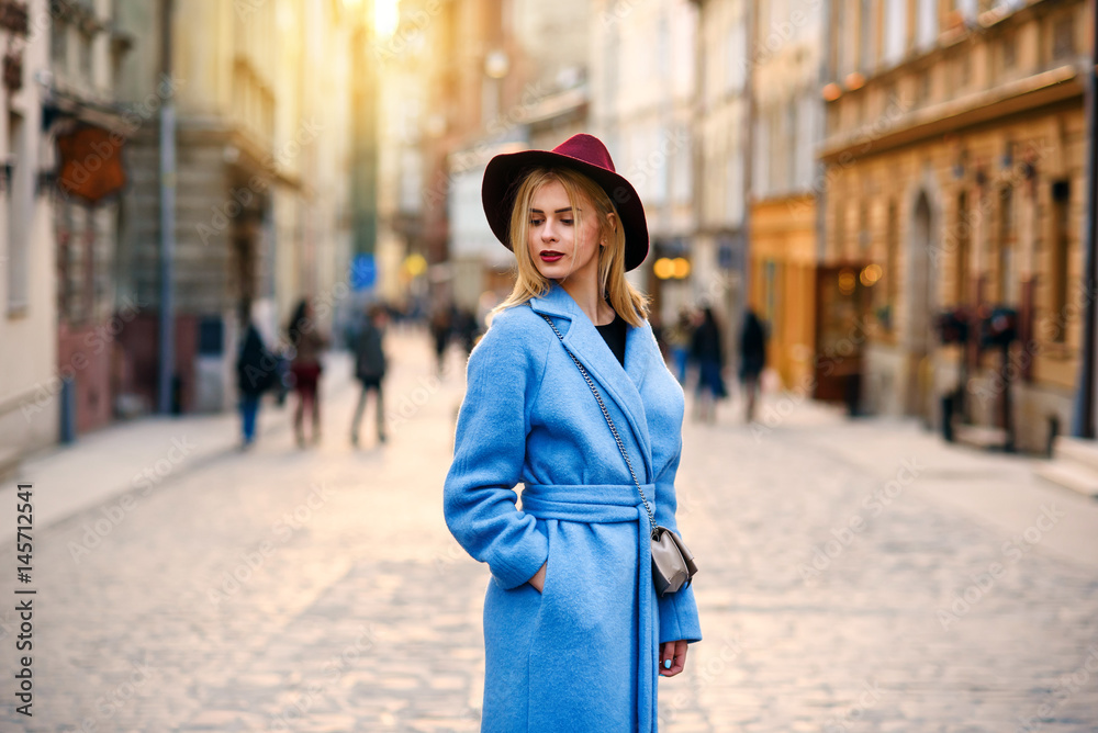 Young beautiful smiling girl in a blue coat and burgundy hat walking down the street on sunset