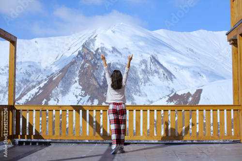 Happy healthy brunette woman in pajama back view stretching on balcony overlooking a mountain snow peak, sunny morning. Vacation in resort hotel. Simple lifestyle people in cozy relaxing apres ski photo