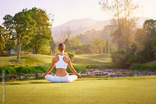 Yoga at park with view of the mountains, with sunlight. Young woman in lotus pose sitting on green grass. Concept of calm and meditation.