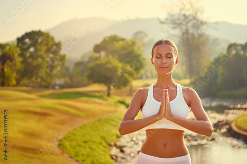 Yoga at park with view of the mountains and lake. Young woman with clasped hands. Concept of calm and meditation.