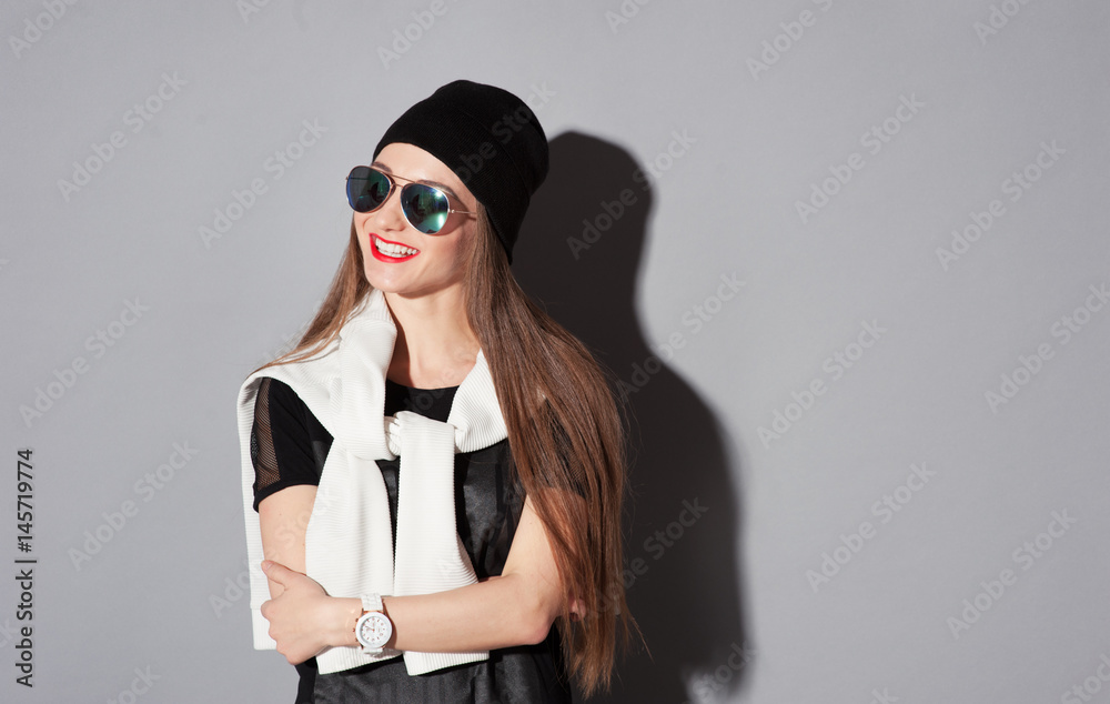 Youth and fashion. Be trendy! Attractive young smiling woman in hat and sunglasses standing against grey background.