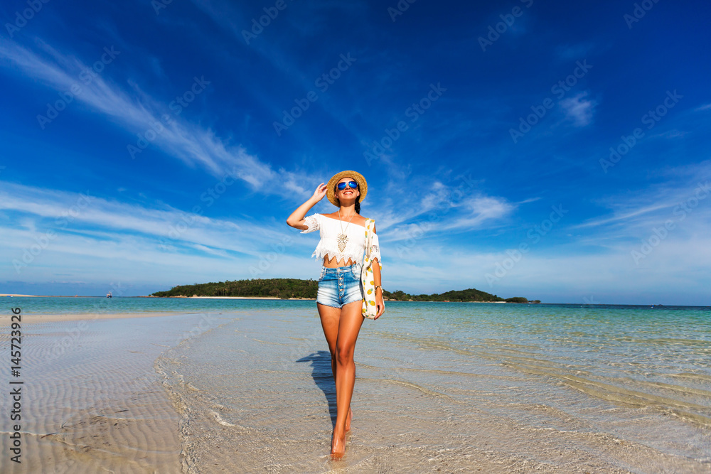 young cute happy girl on the beach, ocean. Dressed in denim shorts, top, straw hat, sunglasses, a bag with pineapple, wide angle photo.