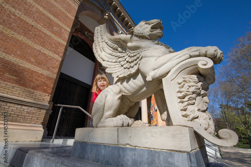 three years old blonde child saluting, leaning on winged lion statue, outside Palace of Velazquez (from year 1883 by architect Ricardo Velazquez Bosco), in public park El Retiro, Madrid, Spain
