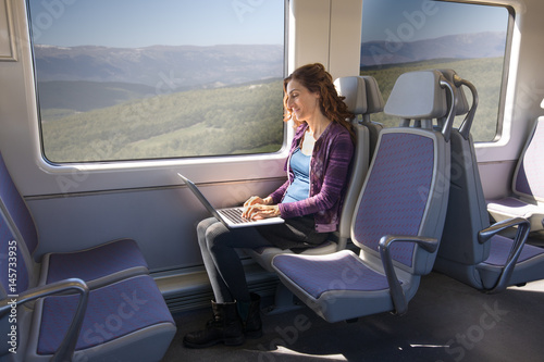 brown hair smiling woman dressed in purple and blue, sitting traveling by train typing in keyword of laptop computer leaning on her legs
