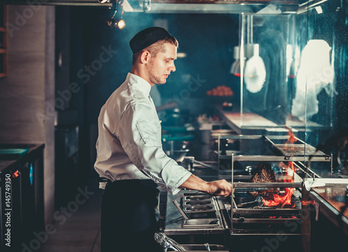 Food concept. Young handsome chef in white uniform monitors the degree of roasting and turns meat with the forceps in interior of restaurant kitchen. Preparing traditional beef steak on barbecue oven.