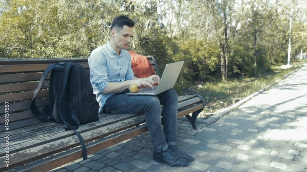 Young man in shirt siting on the bench in green park and typing on his laptop and eating apple Young man in shirt siting on the bench in green park and typing on his laptop and eating apple