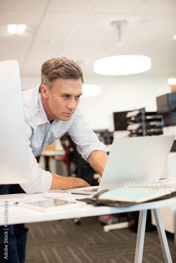 Serious businessman looking at laptop while leaning on desk