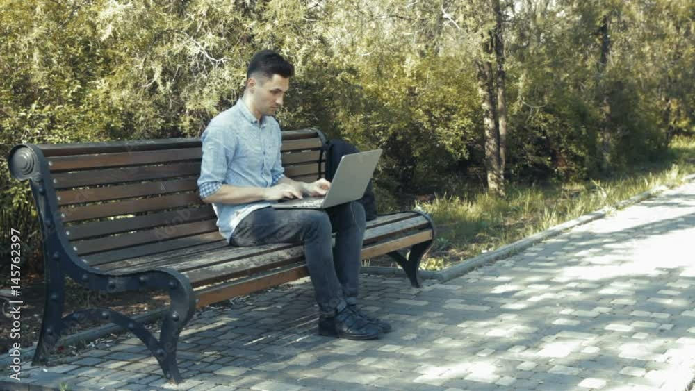 Young man in shirt siting on the bench in green park and typing on his laptop Young man in shirt siting on the bench in green park and typing on his laptop