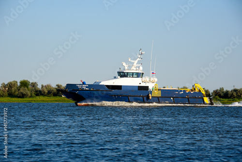 Blue and white ship moving in river, with green plants on background