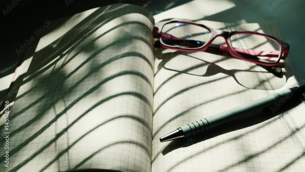 The notepad is open near the window. There are reading glasses and a pen. The sun is shining brightly, the shadows run along the paper. New start and start of business, readiness for fresh ideas The notepad is open near the window. There are reading glasses and a pen. The sun is shining brightly, the shadows run along the paper. New start and start of business, readiness for fresh ideas
