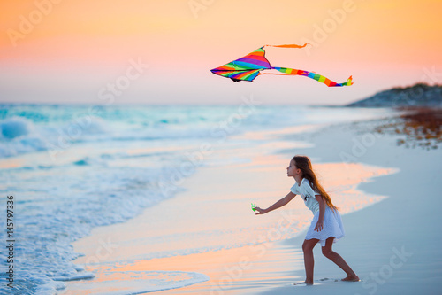 Little running girl with flying kite on tropical beach at sunset. Kids play on ocean shore. Child with beach toys.