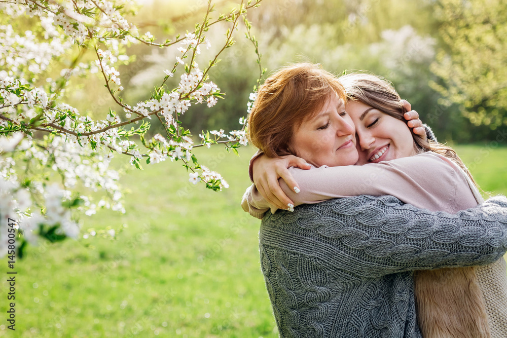 Middle-aged mother and her daughter hugging in blooming garden - obrazy, fototapety, plakaty 