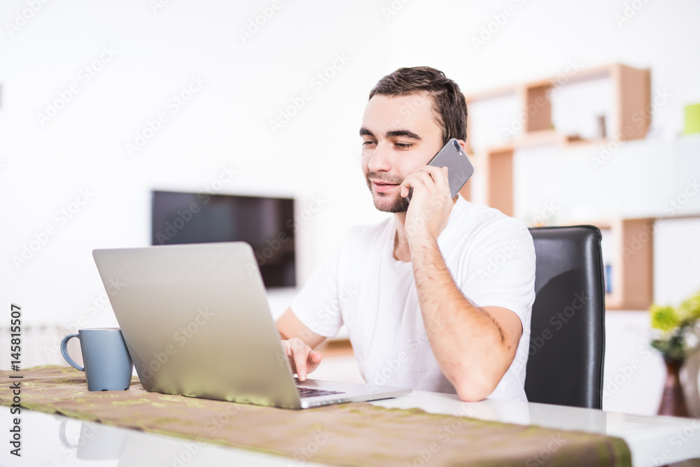 Handsome businessman is talking on the mobile phone and smiling while using a laptop in kitchen Handsome businessman is talking on the mobile phone and smiling while using a laptop in kitchen