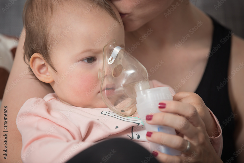 One year old baby girl inhaling from the inhaler, her mother holding her in the arms and comforting the ill baby, kissing her on the head One year old baby girl inhaling from the inhaler, her mother holding her in the arms and comforting the ill baby, kissing her on the head