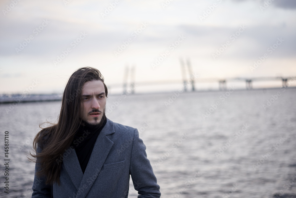 Portrait of concentrated Stylish man with long hair look away. Attractive model posing outdoor in autumn or spring time at seaside of big city. Copy space for a promotion text or advertising Portrait of concentrated Stylish man with long hair look away. Attractive model posing outdoor in autumn or spring time at seaside of big city. Copy space for a promotion text or advertising