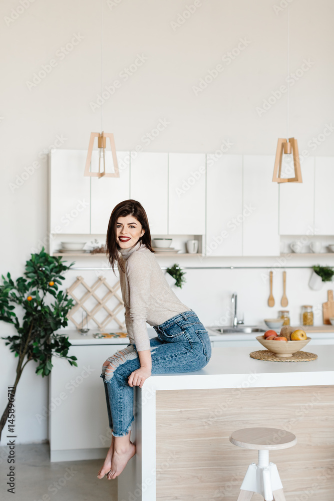 girl sitting on the kitchen table. Bright, white kitchen. Happy smiling girl in the kitchen. Kitchen girl sitting on the kitchen table. Bright, white kitchen. Happy smiling girl in the kitchen. Kitchen
