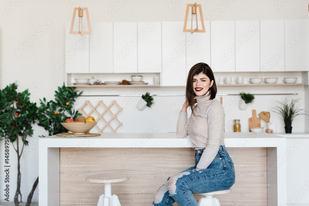 girl standing near the kitchen table in a high chair. Bright, white kitchen. Happy smiling girl in the kitchen. Girl sitting on a chair. Kitchen girl standing near the kitchen table in a high chair. Bright, white kitchen. Happy smiling girl in the kitchen. Girl sitting on a chair. Kitchen