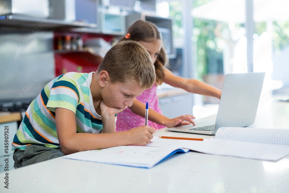 Boy doing his homework while girl using laptop in kitchen Boy doing his homework while girl using laptop in kitchen
