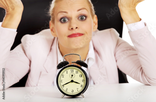 Surprised business woman lsitting behind the desk with alarm clock photo