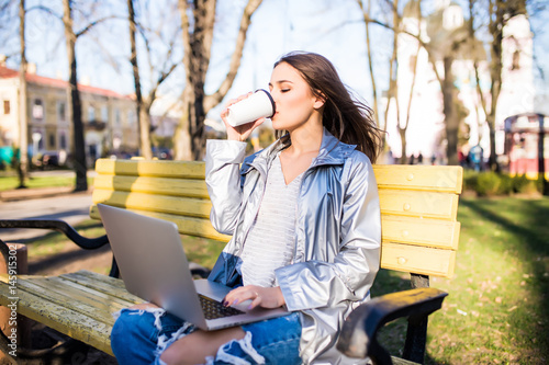 Young woman drink a cup of coffee while using a laptop in a park.