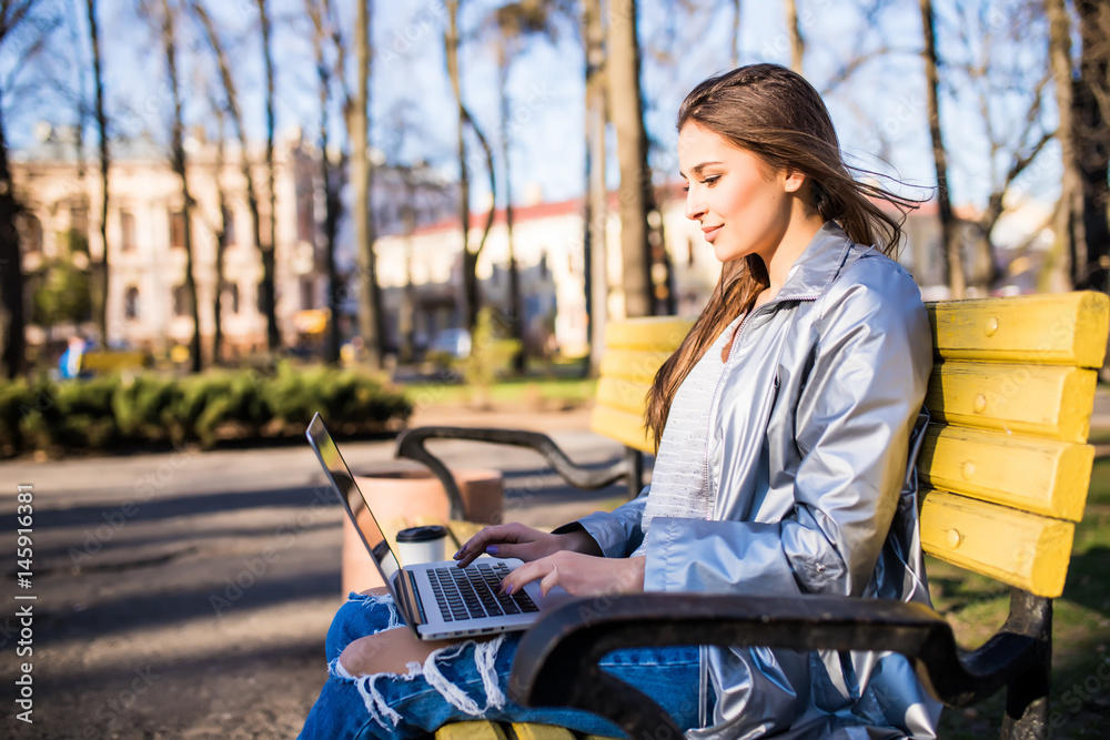 Young pretty brunette woman is sitting on the bench in city and working with laptop.