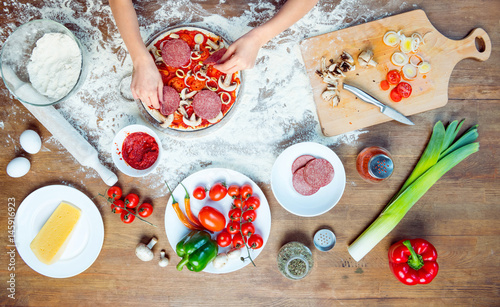 top view of child making pizza with pizza ingredients, tomatoes, salami and mushrooms on wooden tabletop