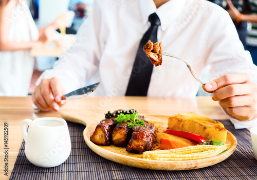 Close up of woman hand holding fork while eating rib steak on wooden tray at restaurant.