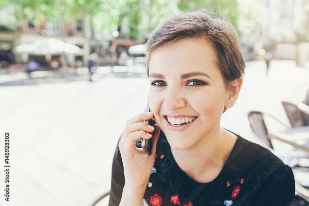 Close-up portrait of attractive young Caucasian woman talking on mobile phone at outdoor cafe. Girl talking by mobile phone, smiling. Sunny day. Outdoor photo. Close-up portrait of attractive young Caucasian woman talking on mobile phone at outdoor cafe. Girl talking by mobile phone, smiling. Sunny day. Outdoor photo.