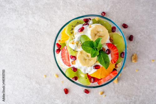 Fresh fruit salad with yogurt and walnuts in glass bowl on stone background.