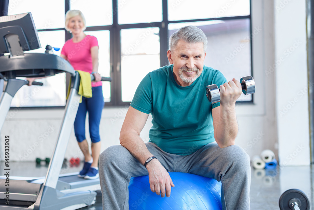 senior sportsman sitting on fitness ball and training with dumbbell, sportswoman on treadmill behind. senior fitness class concept senior sportsman sitting on fitness ball and training with dumbbell, sportswoman on treadmill behind. senior fitness class concept