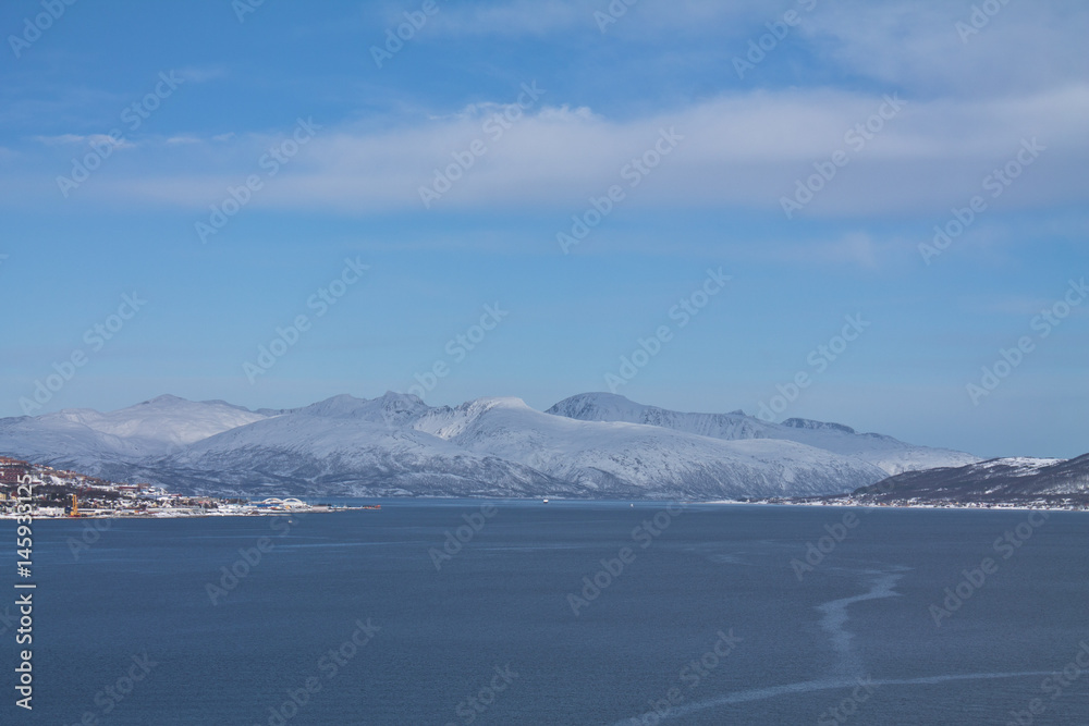 view from boat on Tromso landscape with snowy mountains and city harbor in sunny blue sky, Norway
