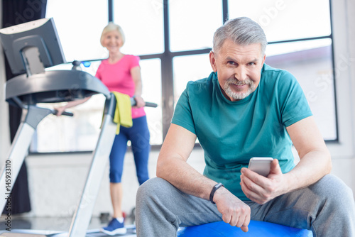senior sportsman sitting on fitness ball and using smartphone, sportswoman on treadmill behind. senior fitness class concept