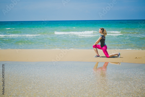 young mother stretching legs with lunge hamstring stretch exercise leg stretches. Fitness female athlete relaxing on beach doing a warm-up before her strength training cardio workout.