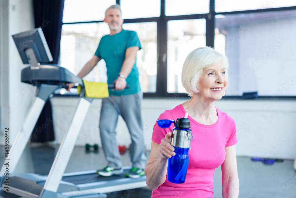 Fototapeta premium Bearded man exercising on treadmill while smiling woman drinking water in fitness class
