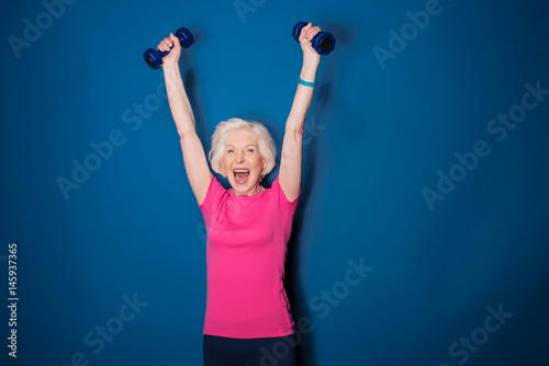 Excited senior fitness woman training with dumbbells isolated on blue