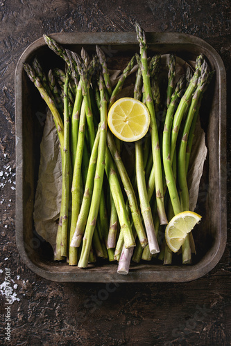 Bundle of young raw uncooked organic green asparagus with sliced lemon and sea salt in old oven tray over brown texture background. Top view. Healthy eating