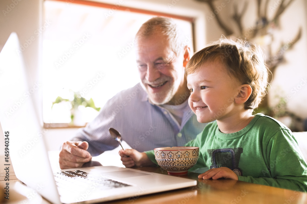Grandfather and grandson sitting at table, eating and using laptop Grandfather and grandson sitting at table, eating and using laptop