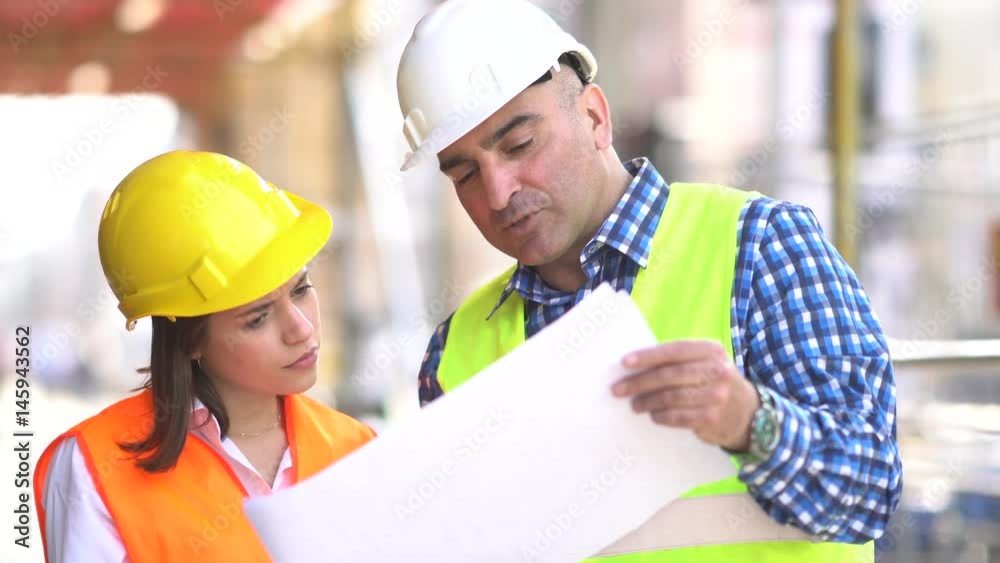 Male and female construction engineers holding plans and inspecting the construction site among scaffolding Male and female construction engineers holding plans and inspecting the construction site among scaffolding
