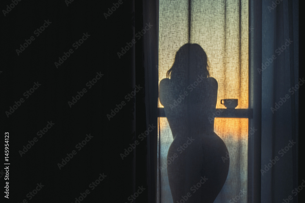 A naked girl is standing by the window behind the curtain and drinking coffee at dawn