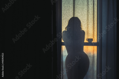 A naked girl is standing by the window behind the curtain and drinking coffee at dawn