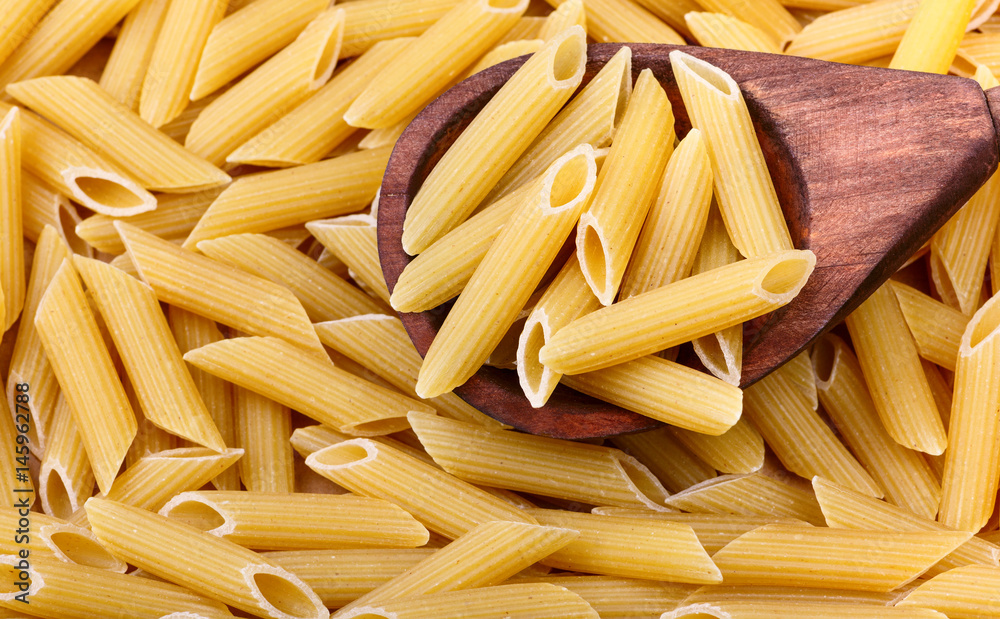 Raw penne pasta in the wooden spoon is on the foreground. Pasta on the background Raw penne pasta in the wooden spoon is on the foreground. Pasta on the background