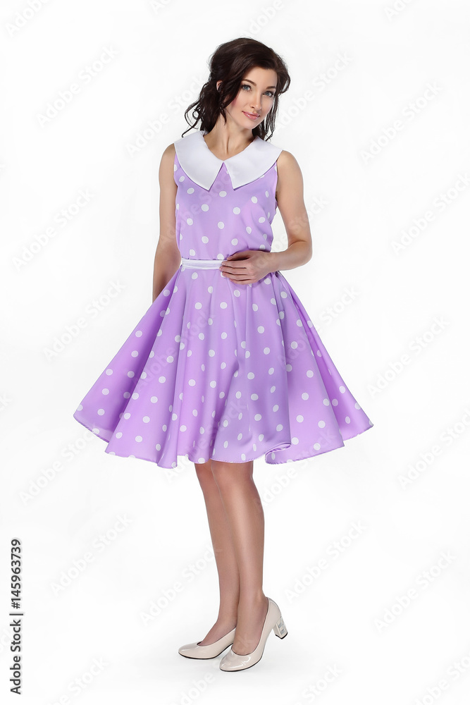 Charming girl in purple dress with polka dots turns around looking at camera on white background