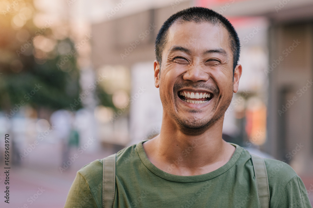Asian man laughing while standing on a city street - obrazy, fototapety, plakaty 