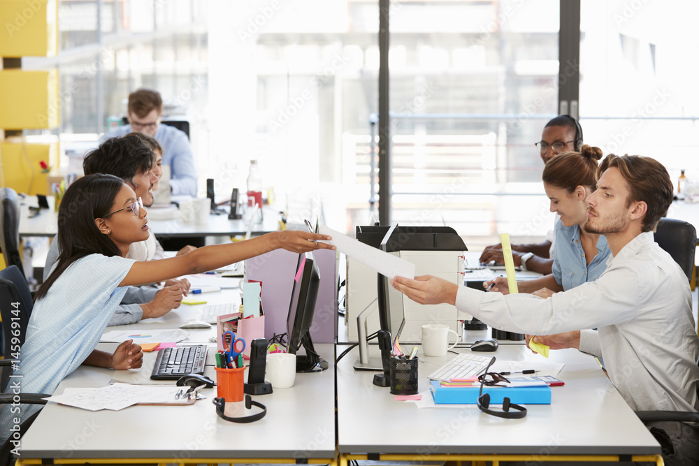 Woman passes document across desk to man in a busy office Woman passes document across desk to man in a busy office