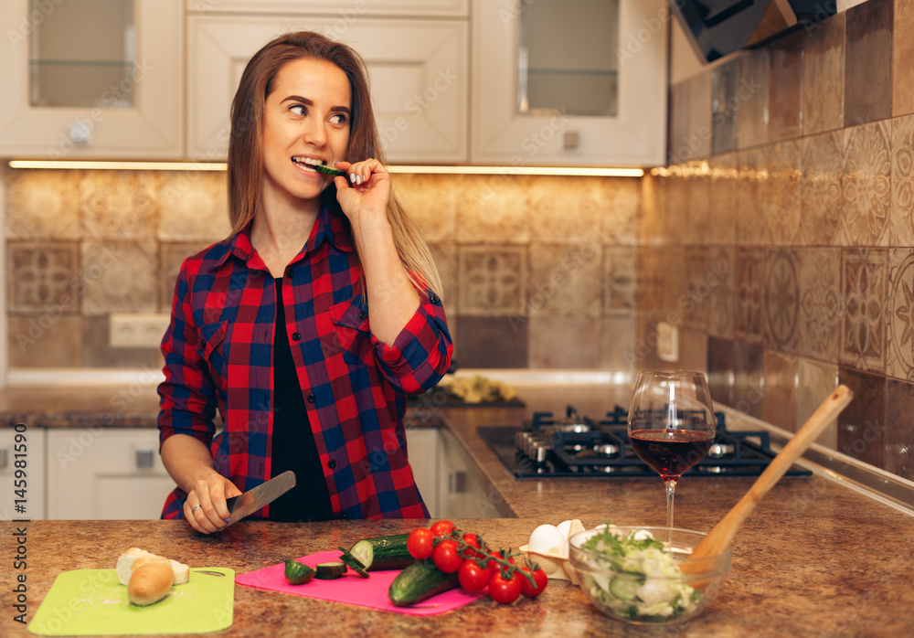 Happy young woman eating slice of cucumber in the kitchen. Woman cutting vegetables for salad.