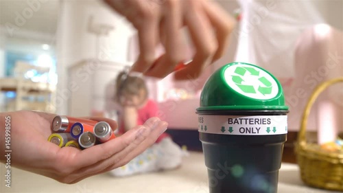Female hands putting used batteries into special recycling box at home. Child playing on the background. Separating Domestic waste. Batteries Only.
