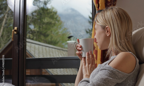 Happy smiling woman drinking hot tea by the window of country house with mountain view. Cozy lifestyle