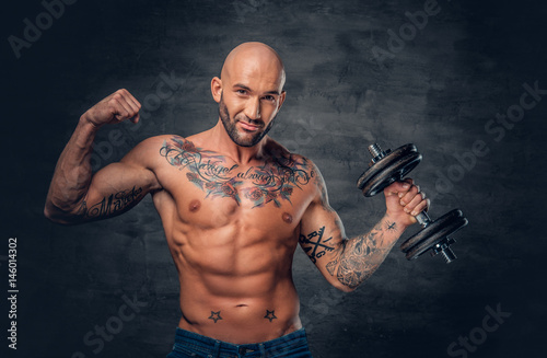 Shaved head sporty male with tattoos on his chest and arms holds dumbbell. © Fxquadro