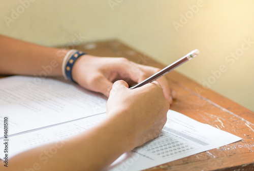 students hand holding pencil fill in Exam carbon paper sheet or test paper on old wood desk in classroom at high school Thailand