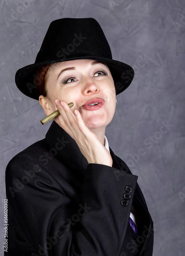 Young woman in manly style with mini cigar on gray background, girl in man's suit and tie, white shirt and hat